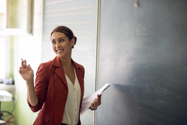 The young female teacher standing in front of the blackboard and showing kids how to solve math problem and multiply. She is holding papers in one hand and white chalk in other. Teacher is asking class do they know correct answer. Back to school concept. Preparing for final exam.