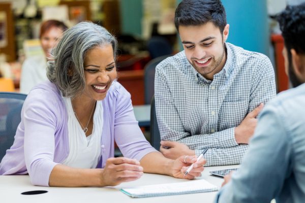 A smiling senior woman sits at a table in her university library with two male classmates as they study class notes.